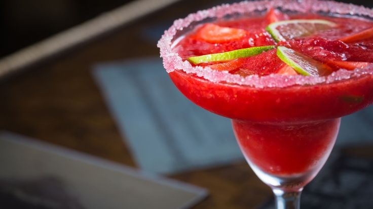 A vibrant red frozen Strawberry Margarita in a salt-rimmed glass, garnished with lime slices. It sits on a dark wooden table, evoking a refreshing and festive mood.